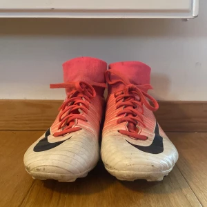 Nike Mercurial rosa fotbollsskor strl 36 - Snygga Nike Mercurial fotbollsskor i rosa och vitt med svarta detaljer. Skorna har högt skaft i stretchigt material, snörning och klassisk Nike-logga på sidan. Perfekta för dig som vill sticka ut på planen!