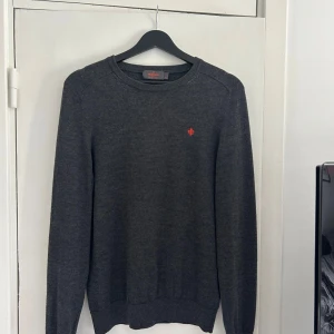 Mörkgrå tröja Morris - Mörkgrå tröja från Morris med rund halsringning och diskret röd logga broderad på bröstet. Klassisk modell med långa ärmar och ribbade muddar. Perfekt för lager-på-lager och enkel att matcha med jeans eller chinos. Storlek M men passar även S