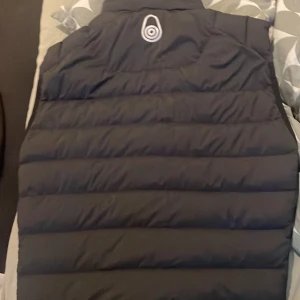 Svart väst från Sail Racing - Säljer en svart dunväst från Sail Racing med hög krage och diskret logga på bröstet. Västen har två fickor med dragkedja och quiltad design. Perfekt för lager-på-lager och snygg till både hoodie och t-shirt.  Normal pris 1000kr