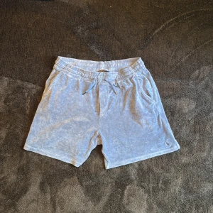 Ljusblå frottéshorts Polo Ralph Lauren - Ljusblå shorts från Polo Ralph Lauren i mjuk frotté med snörning i midjan och broderad logga på benet. Shortsen har sidofickor och en bakficka, perfekt för chill dagar eller när du vill ha en avslappnad look.