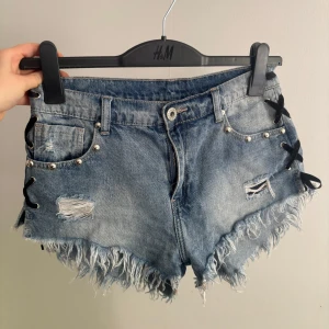 Jeansshorts med nitar och snörning - Snygga blå jeansshorts med slitna detaljer, fransig kant och coola nitar längs fickorna och bak. Sidorna har svart snörning. Står storlek L men skulle säga xs/s