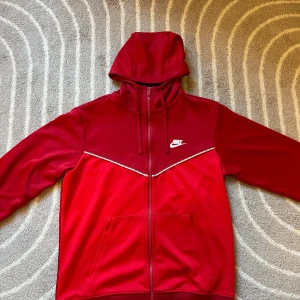 Röd Nike zip-hoodie med logga - Röd hoodie från Nike med dragkedja, huva och snörning. Jackan har vita Nike-loggor längs en diagonal detalj på bröstet och en stor vit logga på vänster sida. Tillverkad i mjuk polyester och har ribbade muddar vid ärmsluten. Perfekt för en sportig look.