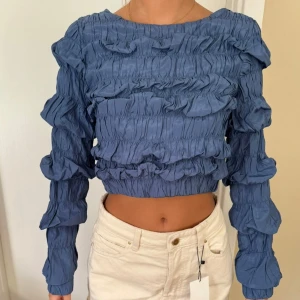 Blå volangblus från Nakd - Supersnygg blå blus från nakd med volangdetaljer och rynkade partier över hela toppen. Långärmad och croppad modell med rund halsringning. Perfekt för dig som vill sticka ut med en cool och trendig look. I storlek xs men passar även s , då det finns mycket stretch i den 