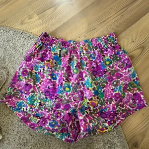 Färgglada blommiga shorts - Säljer ett par loose-fit shorts med elastisk midja och färgstarkt blommönster i lila, blått, grönt och gult. Perfekta för varma dagar när du vill sticka ut. Tunt och mjukt tyg som känns skönt mot huden.