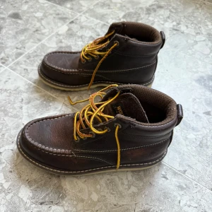 Bruna Wolverine kängor i skinn - Bruna kängor från Wolverine i robust skinn med kontrastsömmar och gula snören. Skorna har en grov, ljus sula och klassisk rund tå. Perfekta för dig som gillar en tidlös och rejäl look med outdoor-vibe.