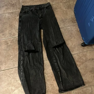 Svarta wide jeans med hål på knäna - Säljer ett par svarta jeans med breda ben och slitna hål över båda knäna. Jeansen har klassisk femficksdesign, dragkedja och knapp framtill. Snygg tvättad look som ger en cool vibe.