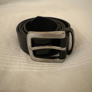 Svart klassiskt skärp i skinn - Snyggt svart bälte i äkta skinn med en stilren, fyrkantig silverspänne. Bältet är slätt utan mönster och har flera hål för justerbar passform. Perfekt för att ge en clean och tidlös look till dina outfits.