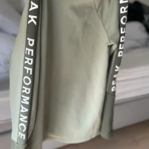 Grön zipjacka Peak Performance - Snygg ljusgrön zipjacka från Peak Performance med svarta detaljer och logga längs ärmarna. Jackan har hel dragkedja, två fickor med dragkedja och sportig look. Perfekt för dig som gillar stilren och bekväm streetwear.