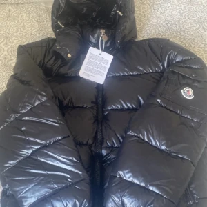  Moncler Maya helt ny  - Säljer en svart, glansig dunjacka från Moncler med huva och dragkedja framtill. Jackan har Moncler-logga på ärmen och ett unikt serietidningsfoder på insidan. Perfekt för kalla dagar och riktigt snygg streetstil.