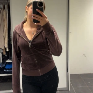 Brun juicycouture kofta - Mysig brun hoodie i velour med dragkedja framtill och huva. Modellen är figurnära med långa ärmar och fickor på magen. Perfekt för en avslappnad stil.