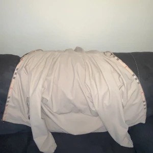 Beige trenchcoat från Burberry - Säljer en klassisk beige trenchcoat från Burberry med det ikoniska rutiga fodret. Jackan har långa ärmar och stilren design, perfekt för dig som vill ha en tidlös look. Storlek M enligt etiketten.
