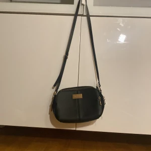 Svart axelväska från Don Donna - Säljer en stilren svart axelväska från Don Donna med justerbart axelband och guldfärgade detaljer. Väskan har dragkedja upptill och en enkel, klassisk design som passar till många outfits.