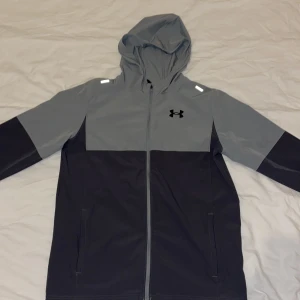  Under Armour vindjacka - Snygg tvåfärgad vindjacka från Under Armour i grått och svart. Jackan har huva, hel dragkedja framtill och två fickor. Perfekt för blåsiga dagar och sportiga outfits. Reflekterande detaljer på axlarna och klassisk logga på bröstet.