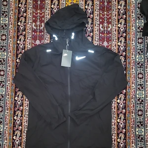 Svart Nike vindjacka med reflexdetaljer - Svart vindjacka från Nike med huva och hel dragkedja framtill. Jackan har flera reflexdetaljer på axlar, ärmar och huva samt meshfoder på insidan. Perfekt för dig som vill ha en sportig look och synas i mörkret. Snygg och funktionell design.