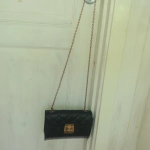 Svart quiltad axelväska med guldkedja - Säljer en svart quiltad axelväska med guldfärgad kedja och låsdetalj framtill. Väskan har en klassisk rektangulär form och är perfekt för att bära det viktigaste. Kedjan ger en snygg kontrast mot det svarta materialet.