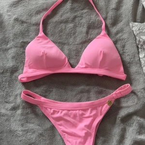 Rosa bikini med trekantsöverdel - Snygg rosa bikini med trekantsöverdel och klassiska bikinitrosor. Överdelen har knyt i nacken och ryggen, och bikinitrosan har en liten gulddetalj på sidan. Perfekt för stranden eller poolen!