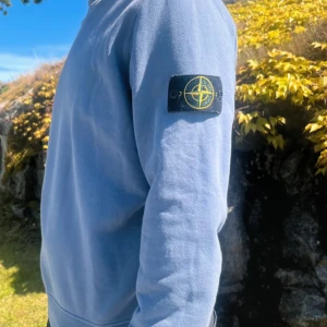 Ljusblå sweatshirt från Stone Island - Säljer en ljusblå sweatshirt från Stone Island med den klassiska loggan på vänster ärm. Tröjan har rund halsringning och långa ärmar. Perfekt för dig som gillar stilrena och exklusiva plagg.