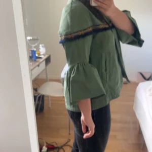Grön jacka med broderad detalj från Zara Basic - Säljer en grön jacka från Zara Basic med broderad kant och vida trekvartsärmar. Jackan har en dekorativ bård i blått och gult längs axlarna och en lös passform. Perfekt för dig som vill ha något unikt och färgglatt i garderoben!