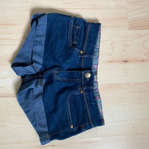 Lågmidjade jeansshorts - Snygga mörkblå jeansshorts med uppvikta ben och klassiska fickor både fram och bak. Insidan av linningen har ett blommigt tyg som detalj. Stl S men skulle säga att de sitter mer som en XS. Säljer då de är för små!