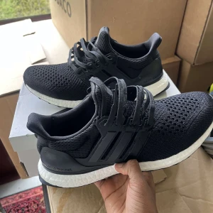 Svarta Adidas Ultraboost sneakers - Säljer ett par svarta Adidas Ultraboost sneakers med vit Boost-sula. Skorna har stickad ovandel, klassisk snörning och tre svarta ränder på sidan. Perfekta för både träning och vardag, riktigt sköna och stilrena.