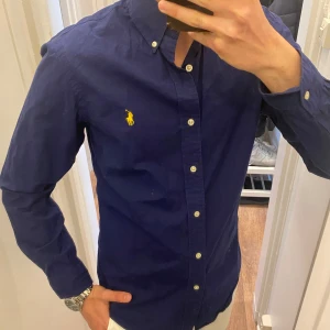 Mörkblå Skjorta från Polo Ralph Lauren - Säljer en klassisk mörkblå skjorta från Ralph Lauren med button-down krage och vita knappar. Skjortan har lång ärm och den ikoniska gula broderade loggan på bröstet. Perfekt för dig som gillar stilrena plagg med snygga detaljer. Storlek Medium. Snabb Leverans!