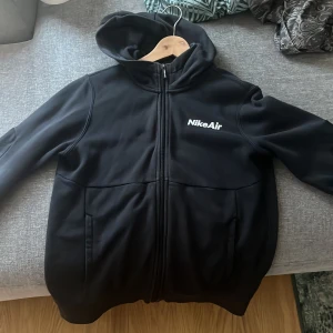 Svart hoodiejacka från Nike Air för barn  - Säljer en svart hoodiejacka från Nike Air med dragkedja framtill och två fickor. Jackan har huva och Nike Air-logga på bröstet. Perfekt för en sportig och avslappnad stil.
