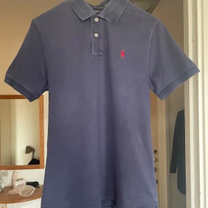Mörkblå pikétröja från Polo Ralph Lauren - Säljer en mörkblå pikétröja från Polo Ralph Lauren med klassisk krage och knäppning framtill. Tröjan har röd broderad logga på bröstet och är i slim fit-modell. Perfekt för en stilren och avslappnad look. Sjukt fräsch till sommaren med ett par shorts! Var uppmärksam på att piken är L för 18-20 år.