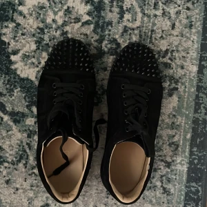 Svarta sneakers med nitar och röd sula - Säljer ett par svarta sneakers med coola svarta nitar på tån och röd sula undertill. Skorna har snörning och är tillverkade i mocka med en platt sula. Perfekta för dig som vill sticka ut med din stil.