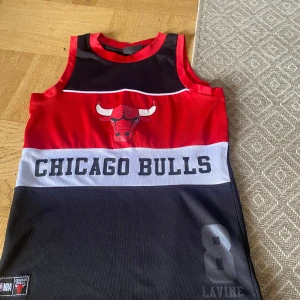 Chicago Bulls basketlinne NBA - Säljer ett Chicago Bulls basketlinne med tryck av lagets logga och texten 'CHICAGO BULLS' på framsidan. Linnet har nummer 8 och namnet LaVine tryckt nedtill. Färgerna är svart, röd och vit. Perfekt för basketfans!