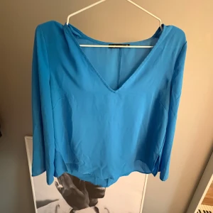 Blå v-ringad blus från GinaTricot  - Säljer en blå blus från GinaTricot med v-ringning och långa ärmar. Blusen har en lös passform och är tillverkad i ett tunt, lätt material som ger ett snyggt fall. Perfekt att styla med jeans eller kjol.