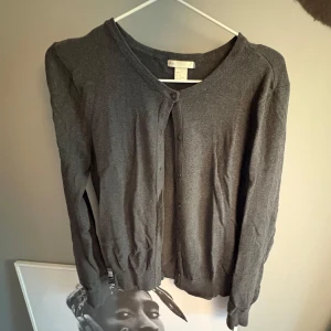 Mörkgrå kofta från H&M - Mörkgrå klassisk kofta från H&M med rund halsringning och knäppning framtill. Långärmad modell i mjukt material, perfekt för lager-på-lager. Enkel och stilren design som passar till det mesta.