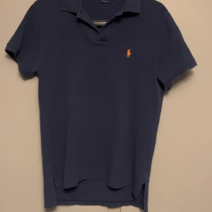 Blå pikétröja från Polo Ralph Lauren - Säljer en klassisk blå pikétröja från Polo Ralph Lauren med orange broderad logga på bröstet. Tröjan har korta ärmar, krage och knappar vid halsen. Perfekt för en stilren och avslappnad look.