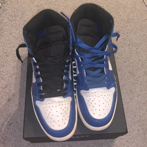 Nike Air Jordans blå och vita high-top sneakers - Säljer ett par Nike Air Jordans high-top sneakers i blått, vitt och svart. Skorna har klassisk rund tå, perforerad tåbox och snörning. Ena skon har svarta skosnören och den andra blå. Perfekta för dig som gillar streetwear och vill sticka ut.