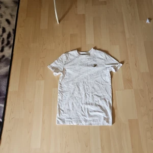 Vit t-shirt från Lyle & Scott Junior - Vit t-shirt från Lyle & Scott Junior med klassisk passform och korta ärmar. På bröstet sitter den ikoniska broderade gula örnen. Tillverkad i mjuk bomull, perfekt för en clean och stilren look. För 10/11 år