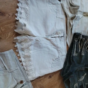 Vita shorts med spetsdetaljer - Säljer ett par vita shorts med breda spetskanter längs benen och broderade blommor vid fickorna. Shortsen har normal passform och är perfekta för sommaren. De har bälteshällor och stängs med knapp och dragkedja.