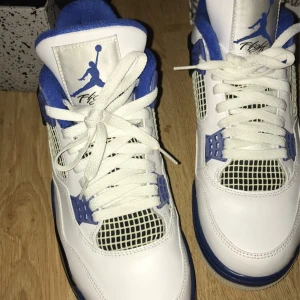 Nike Air Jordan 4 Retro OG 'Military Blue'  Motorsports - Säljer ett par Nike Air Jordan 4 Retro OG 'Military Blue'  Motorsports med vit ovandel i skinn, blå och svarta detaljer samt meshpaneler. Klassisk Jumpman-logga på plösen och platt sula. Skorna har snörning och ikoniska plastdetaljer vid snörningen.