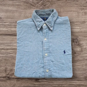 Ljusblå skjorta från Ralph Lauren - Säljer en ljusblå långärmad skjorta från Ralph Lauren med klassisk krage och knappar framtill. Insidan av kragen har ett röd- och blå-rutigt tyg som ger en snygg kontrast. Broderad logga på bröstet. 