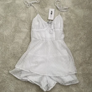 Vit kortklänning från FB Sister - Supersöt vit kortklänning från FB Sister med tunna axelband som knyts, volangig kjol och diskret prickigt mönster i tyget. Klänningen har omlottkänsla, liten rosett framtill och är perfekt för sommarens alla tillfällen. Shorts under klänningen 