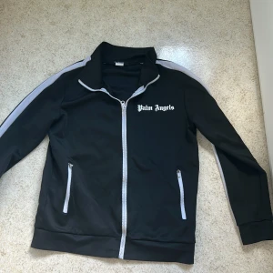 Palm Angels svart trackjacka - Svart trackjacka från Palm Angels med vita ränder längs ärmarna och vit dragkedja. Jackan har hög krage, två fickor med dragkedja och logga tryckt på bröstet. Perfekt för en sportig och trendig streetwear-look.