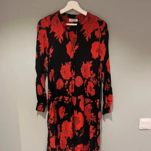 Svart långklänning med röda blommor från Msch - Supersnygg långklänning från Msch i svart med stora röda blommor. Klänningen har långa ärmar, knappar framtill och ett flowigt fall. Perfekt för dig som vill sticka ut med ett coolt blommigt mönster och skön passform. Något mindre i storlek, passar en XS eller mindre S. Ungefär vadlång på mig (1,60cm). 