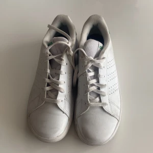 Adidas Advantage JR sneakers - Klassiska Adidas Stan Smith sneakers i vitt skinn med gröna detaljer på hälen. Skorna har perforerade ränder på sidorna, rund tå och snörning framtill. Perfekt för dig som gillar en clean och tidlös look.