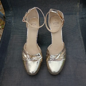 Guldiga klackskor från VOX stl 41 - Säljer ett par glansiga guldiga klackskor från VOX med ormskinnsmönster och ankelrem. Skorna har rund tå och hög klack, perfekta för att sticka ut på festen. Stl 41