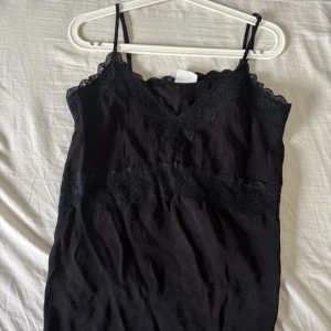 Svart linnetopp med spetsdetaljer - Säljer en svart linnetopp med tunna axelband och v-ringning. Toppen har fina spetsdetaljer längs halsringningen och under bysten, vilket ger en elegant touch. Perfekt att styla till olika outfits!