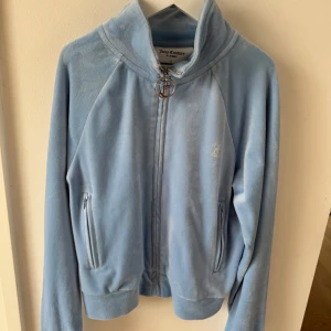 Ljusblå velourkofta från Juicy Couture - Ljusblå velourkofta från Juicy Couture med dragkedja och glittrande Juicy-text på ryggen. Klassisk JC-dragkedja och två fickor framtill. 