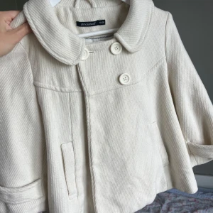 Beige kort kappa från Atmosphere - Säljer en beige kort kappa från Atmosphere med stora knappar framtill och bred krage. Jackan har fickor på sidorna och dekorativ detalj bak. Perfekt för vår och höst.