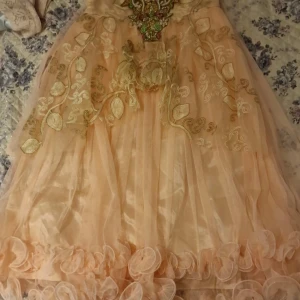 Långklänning med broderier och pärlor - Elegant långklänning i champagnefärg med vackra broderier och pärldetaljer i guld, grönt och rosa. Klänningen har volangdetaljer nertill och ett lager av tunn tyll. Klänning princess vibes. Ingår matchande underbyxor och t-shirt. Det är barnstorlek 36. Den är ej använd.