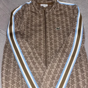 Beige trackjacka med mönster från Lacoste - Snygg trackjacka från Lacoste i beige med geometriskt mönster och bruna, vita och ljusblå ränder längs ärmarna. Jackan har dragkedja och ribbade muddar. Säljer den rätt så billigt då den är slutsåld överallt och knappt använd.