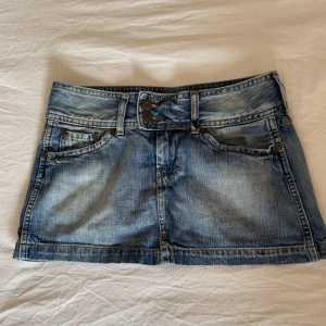 Jeanskjol från Pepe Jeans - Midjemått 35cm Längd 28cm Säljer en klassisk blå jeanskjol från Pepe Jeans med fem fickor, dubbla knappar och bälteshällor. Kjolen är kort och har en lätt tvättad look med slitningar för en avslappnad stil. Perfekt till sneakers eller boots.