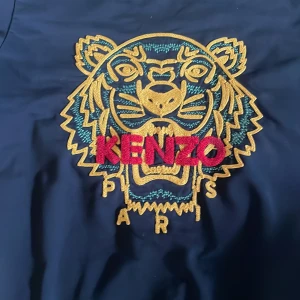 Svart sweatshirt med tigerbrodyr från Kenzo - Säljer en svart sweatshirt från Kenzo med ikonisk tigerbrodyr i gult, grönt och rött framtill. Tröjan har rund halsringning och långa ärmar. Perfekt statement-plagg för dig som gillar streetwear och snygga detaljer.