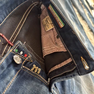 Blå jeans från Jacob Cohen, modell 711 - Säljer ett par snygga blå jeans från Jacob Cohen, modell 711. Jeansen har klassisk femficksdesign, gul kontrastsöm, och en liten italiensk flaggdetalj vid fickan. Raka ben och normal passform, tillverkade i mjukt jeanstyg med lätt tvättad look.